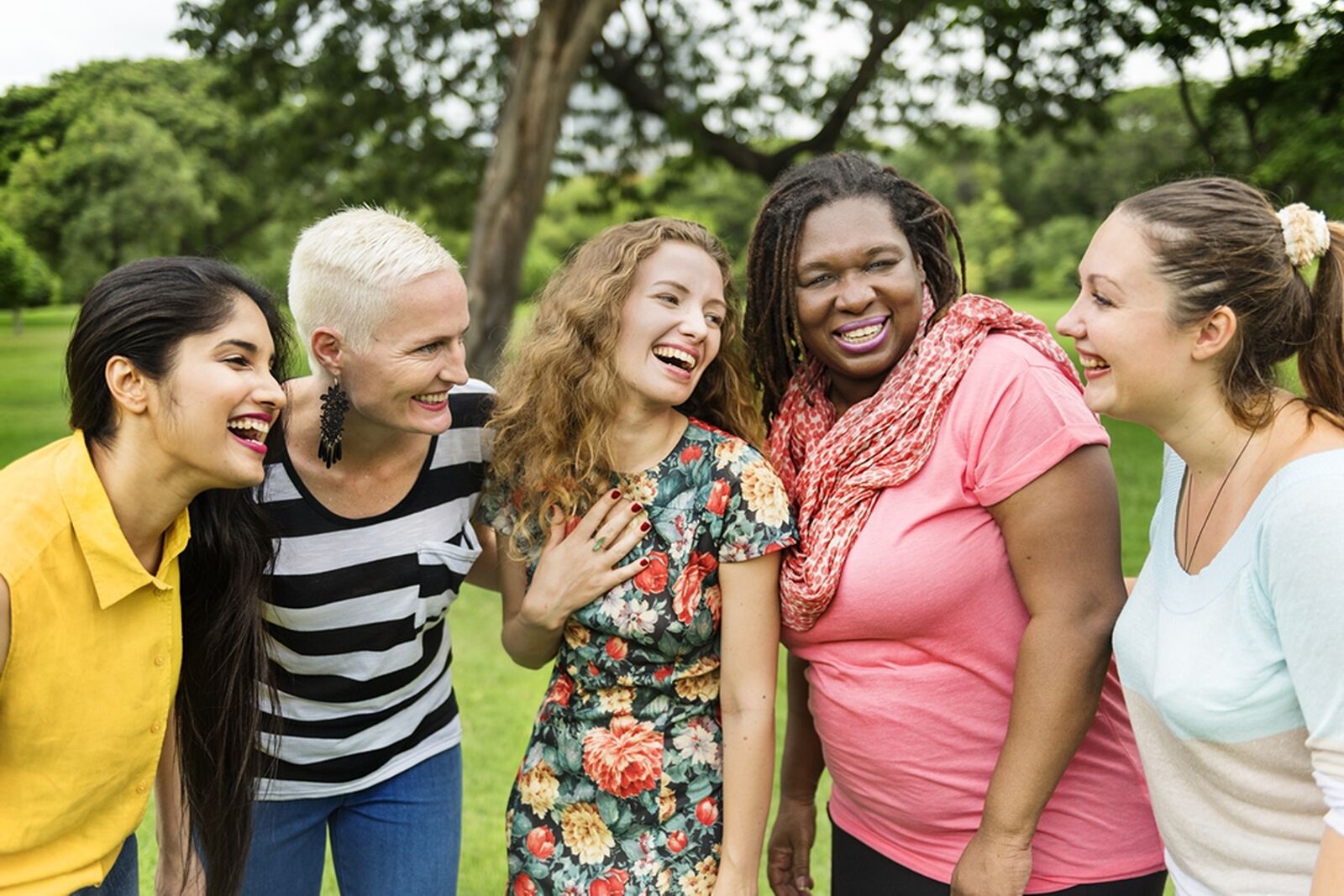 Five women standing together laughing outside in a park