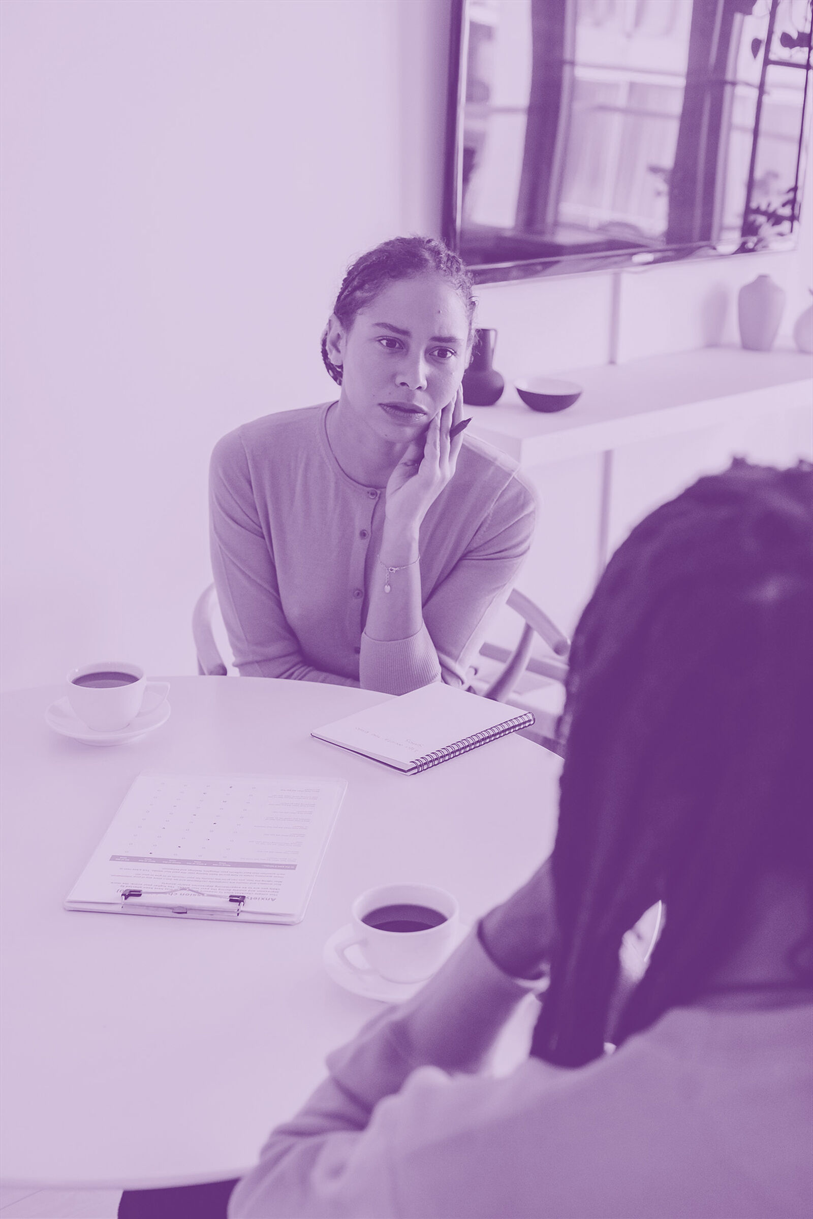 Woman sitting at a table taking notes and talking to person drinking tea purple