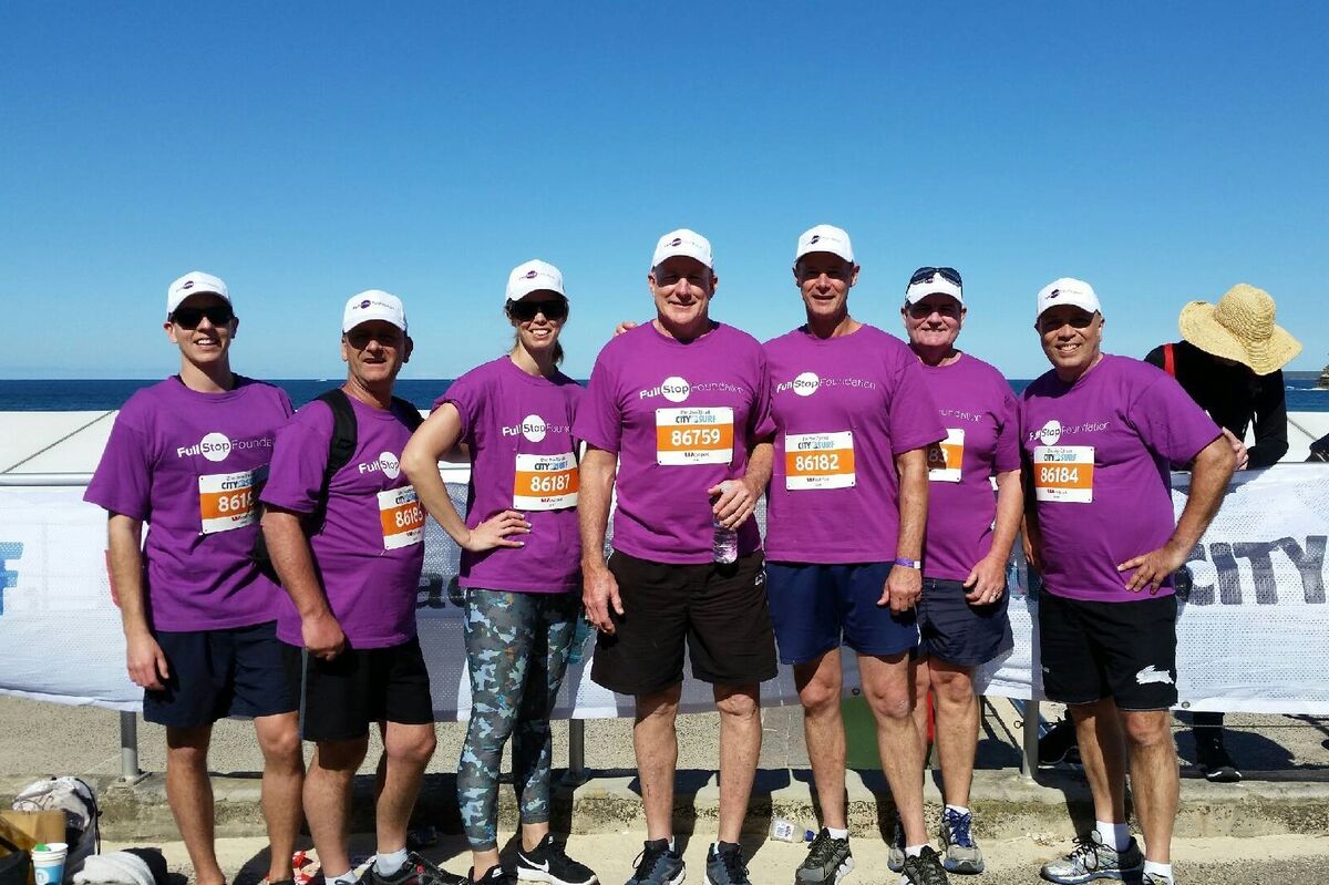 7 people wearing Full Stop Foundation t-shirts stand together smiling near beach at the Sydney City to Surf event