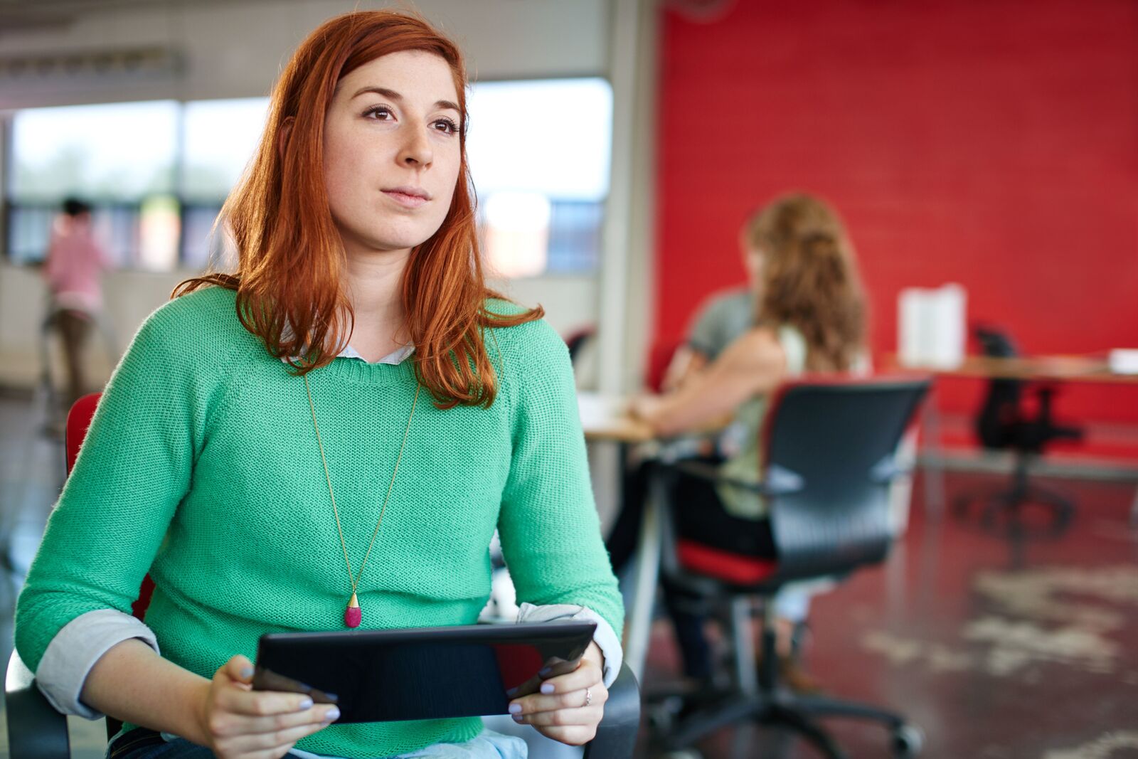 Woman with red hair and a green jumper holding a tablet computer in a workspace.